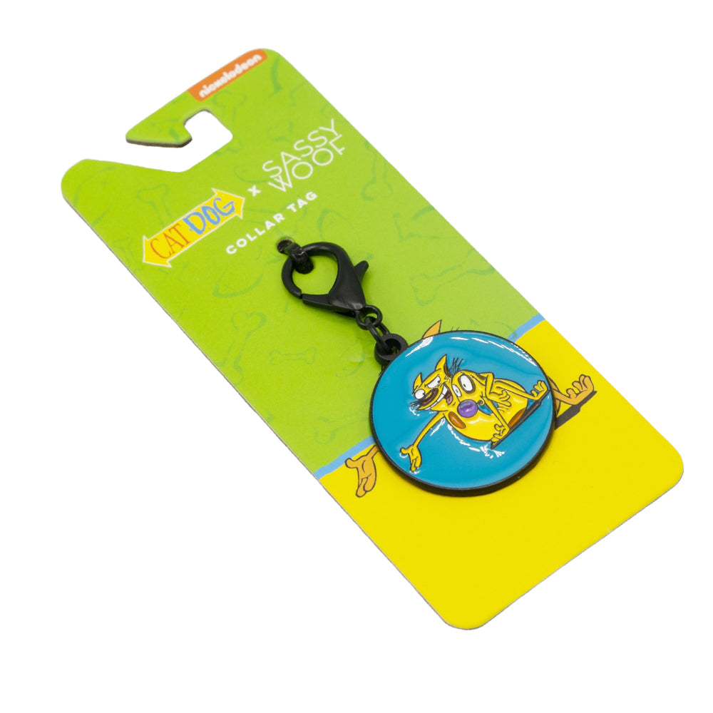 The Sassy Woof Dog Collar Tag - Nickelodeon CatDog™ features the cartoon CatDog on a blue background, attached to yellow Sassy Woof packaging, and is part of the playful CatDog collection with faint character outlines on the back.