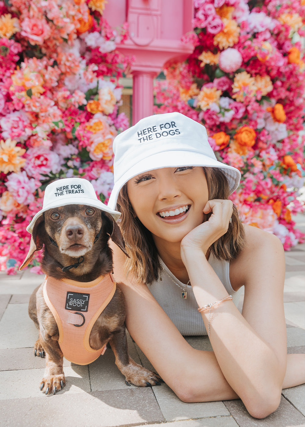 A smiling woman and a brown dog wear matching white bucket hats with text; hers says Here for the dogs, and the dogs says Here for the treats. They pose in front of a vibrant, colorful flower wall.