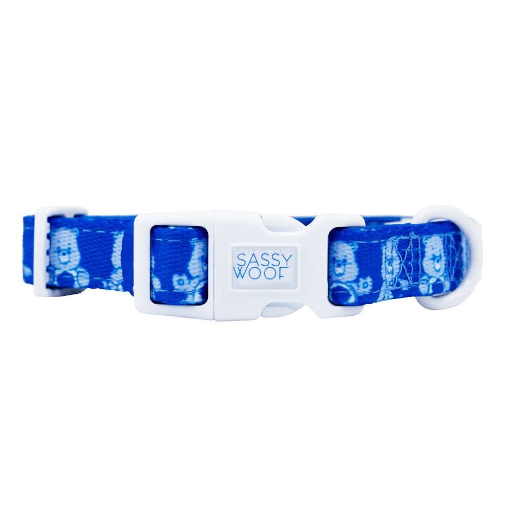 The Sassy Woof Dog Collar - Care Bears™ Grumpy Bear features blue fabric with white cartoon dog faces, a sturdy white plastic "Sassy Woof" buckle, and white metal hardware—perfect to match with your favorite neoprene harness. Shown on a white background.