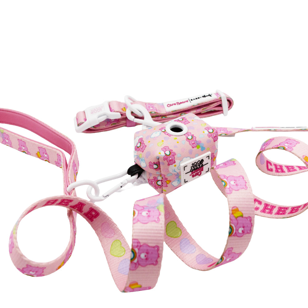 The Sassy Woof Collar Three Piece Bundle - Care Bears™ Cheer Bear features pink harness and leash with playful cartoon bears, heart patterns, a small pouch, and white plastic buckles.