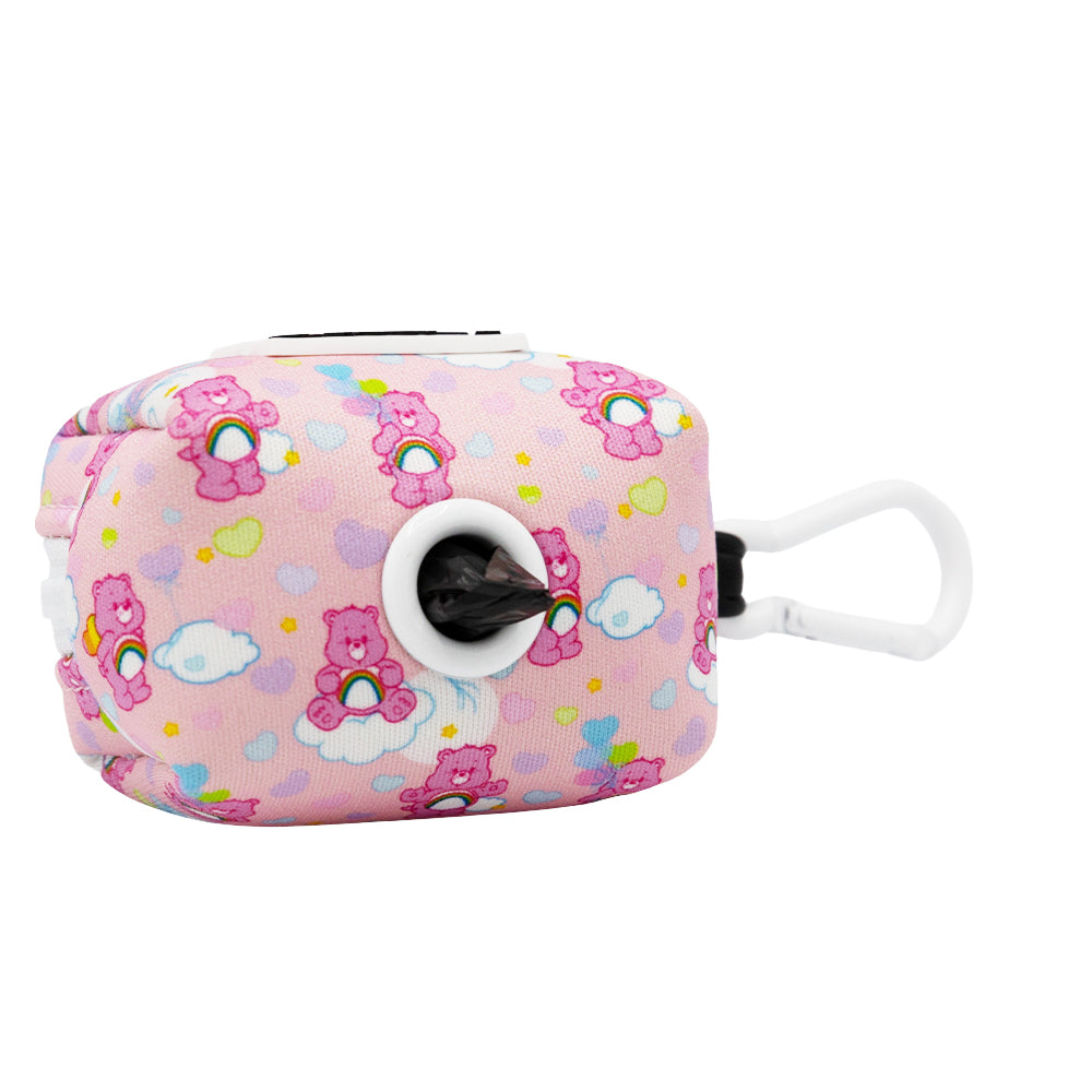 The Sassy Woof Dog Waste Bag Holder - Care Bears™ Cheer Bear is a pink fabric dispenser with hearts, clouds, and Care Bears, plus a white clip for your dog’s harness. Comes with a black bag partially pulled out.
