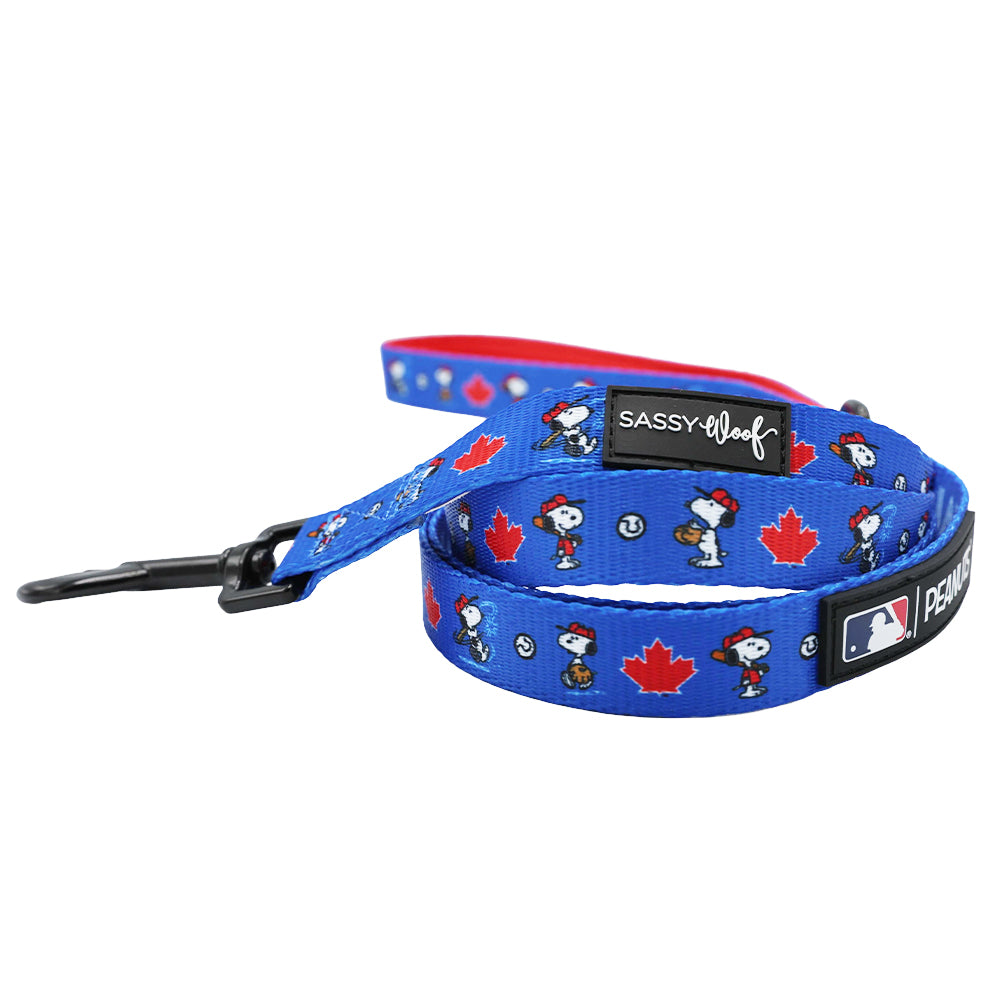 The Sassy Woof Dog Leash - MLB™ x Peanuts™ Toronto Blue Jays™ features blue Snoopy, baseball, and red maple leaf designs, a black plastic clip, Sassy Woof and MLB logos, and a red inner lining with maple leaves.