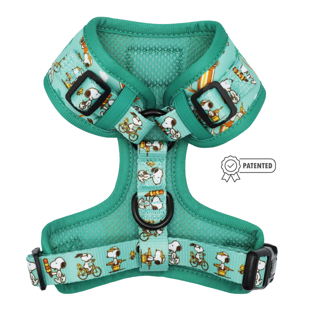 The Sassy Woof Dog Adjustable Harness - Peanuts™ Road Tripping is a teal harness with cartoon Snoopy in action, black buckles, a patent badge icon on the right, and is part of the Puppy Growth Pawtection Plan.