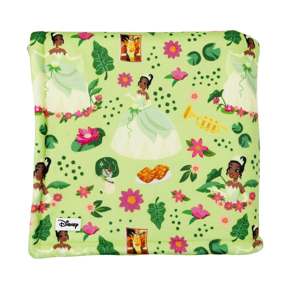 The Sassy Woof Pet Blanket - Disney's Tiana is a soft, light green sherpa dog blanket with Princess Tiana, flowers, leaves, a trumpet, frog, and food illustrations. Features a small white Disney tag on the corner.