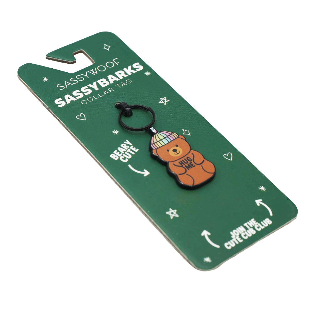 A Sassy Woof Dog Collar Tag - Hug Me Bear package featuring a bear in a striped beanie with "Hug Me" on the tag. Packaging reads "BEARY CUTE" and "JOIN THE CUTE CUB CLUB.