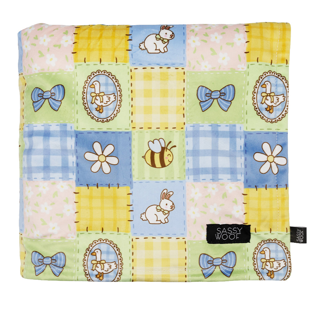 The Sassy Woof Pet Blanket - Patch Perfect features a pastel quilted pattern with rabbits, bees, bows, flowers, and gingham squares plus a black “Sassy Woof” label sewn in one corner.