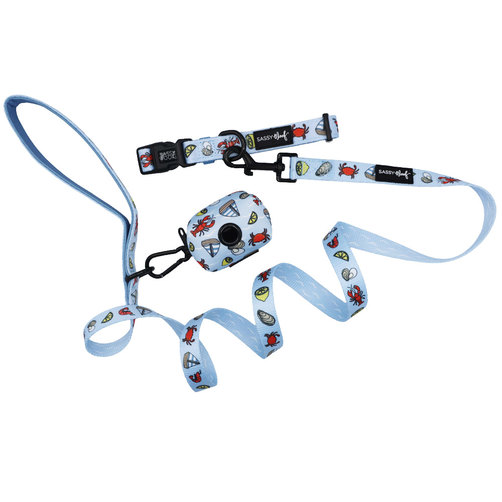 The Sassy Woof Dog Three Piece Bundle - Coastal Cuties features a light blue leash, collar, and matching waste bag holder with fun cartoon crabs, shells, and fish. Set is displayed on a plain white background.