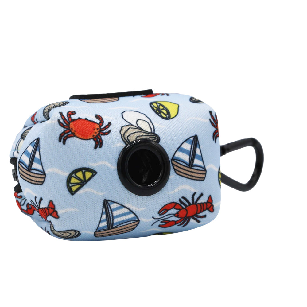 The Sassy Woof Dog Waste Bag Holder - Coastal Cuties is a small, light blue neoprene holder with a black dispenser, decorated with crabs, lime wedges, oysters, and sailboats. Includes a black loop for carrying or hanging.