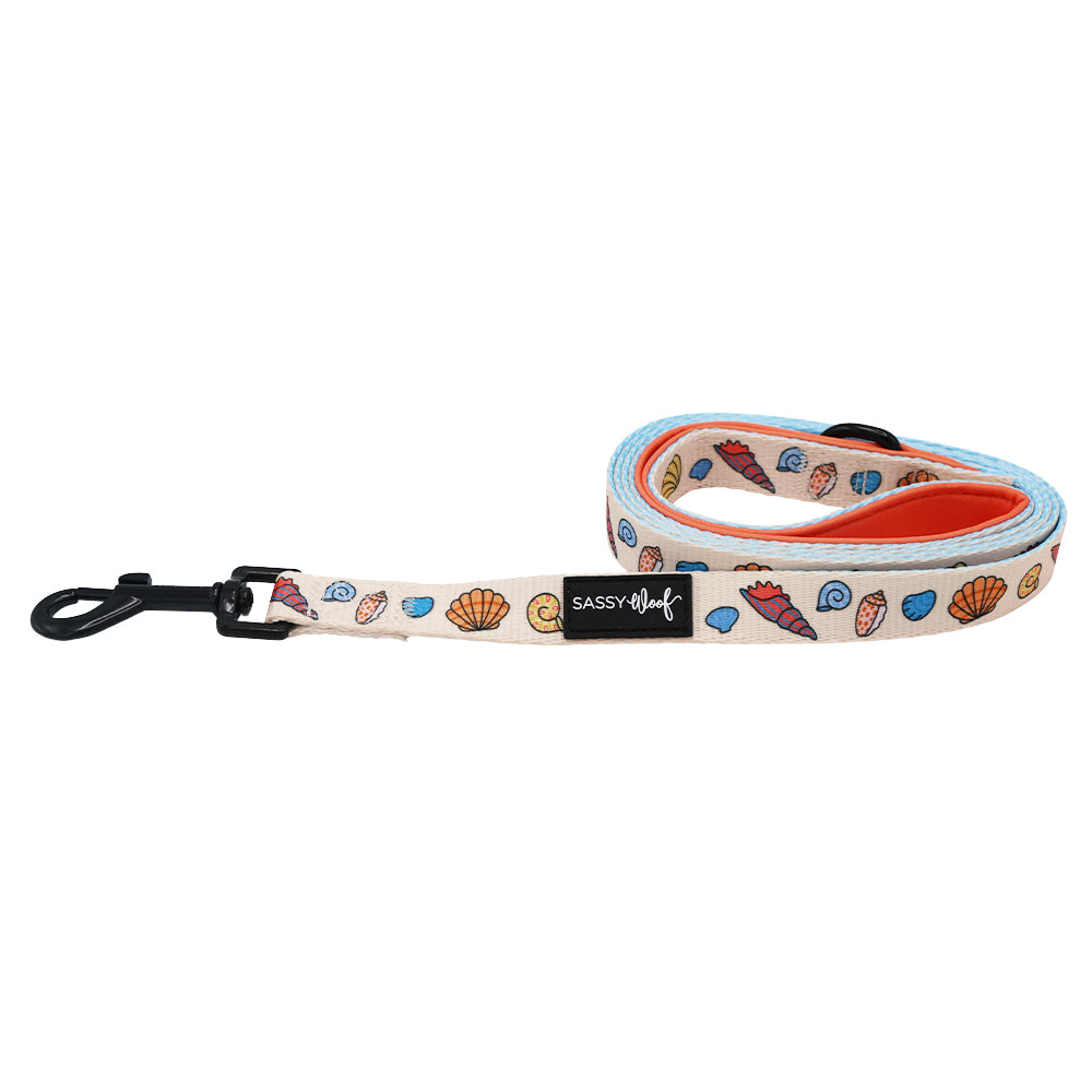 The Sassy Woof "Shell Yeah" dog leash features a beige seashell design with blue trim, orange underside, black metal clasp, and a Sassy Woof label—ideal for beach-loving pups and their owners.