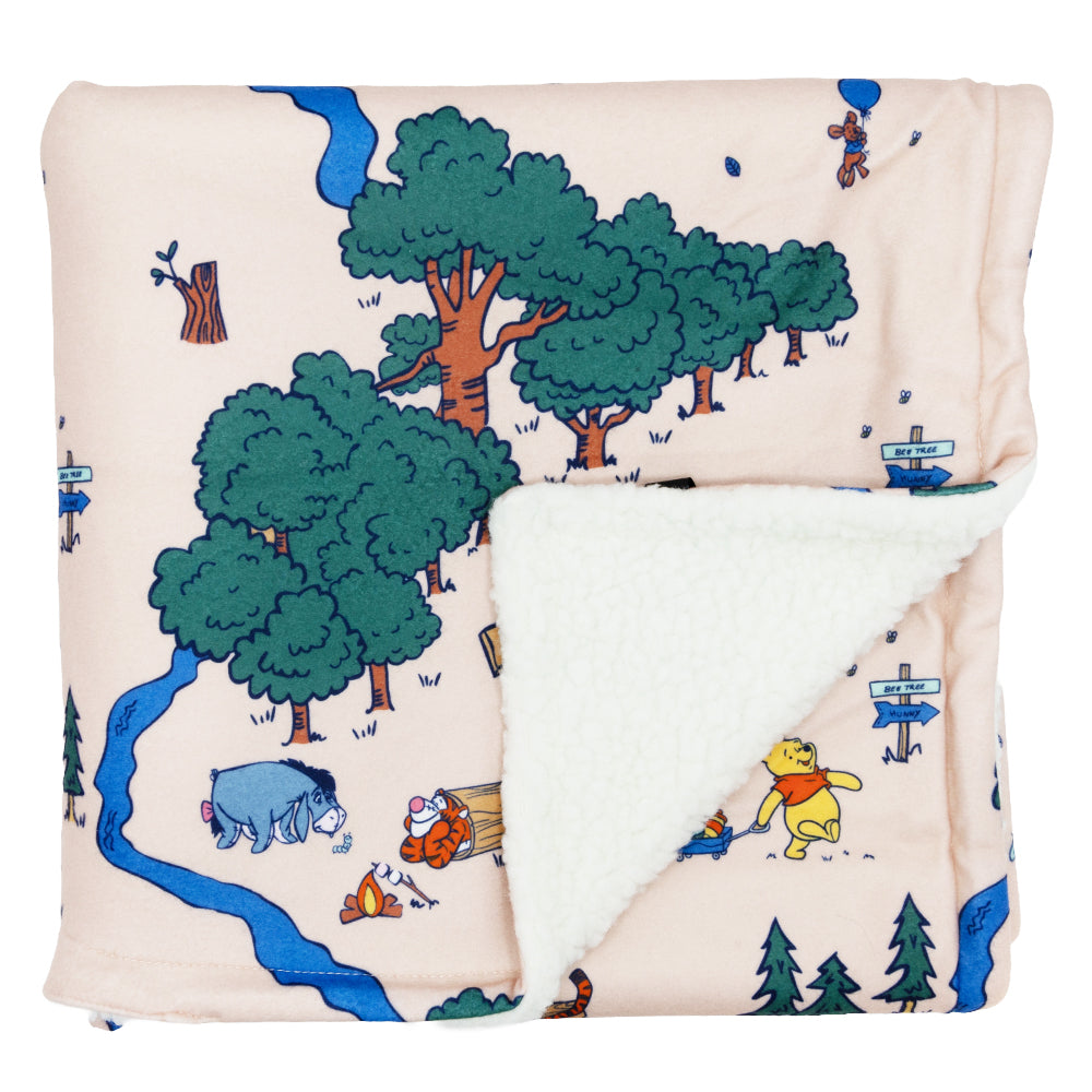 The Sassy Woof Pet Blanket – Disney's Winnie the Pooh features soft fabric with Winnie the Pooh characters and trees on a light background, plus a cozy white sherpa lining revealed on a folded corner.