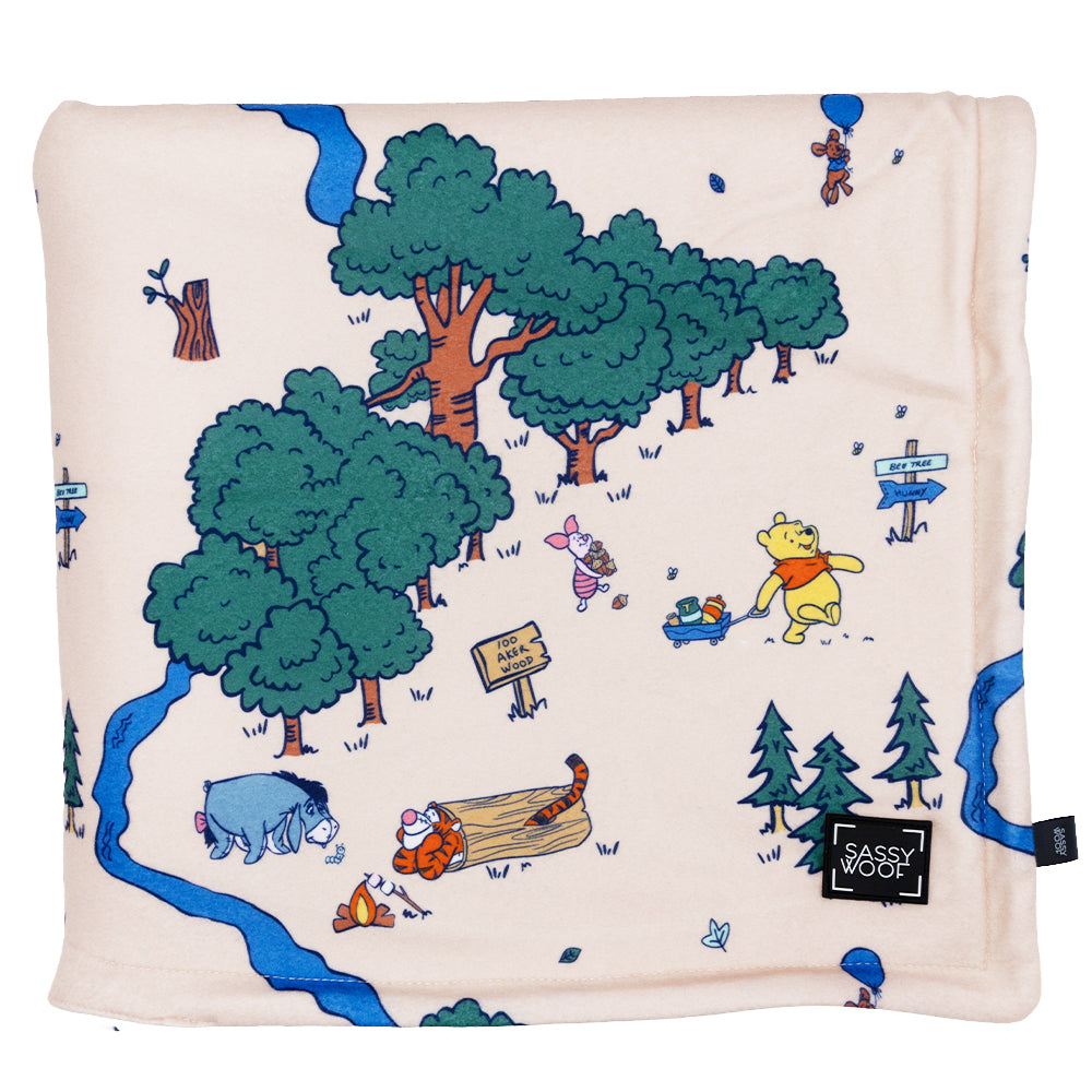 The Sassy Woof Pet Blanket - Disney’s Winnie the Pooh is a cream sherpa blanket with illustrated Hundred Acre Wood characters, trees, signs, and a blue river. It features a black Sassy Woof label.