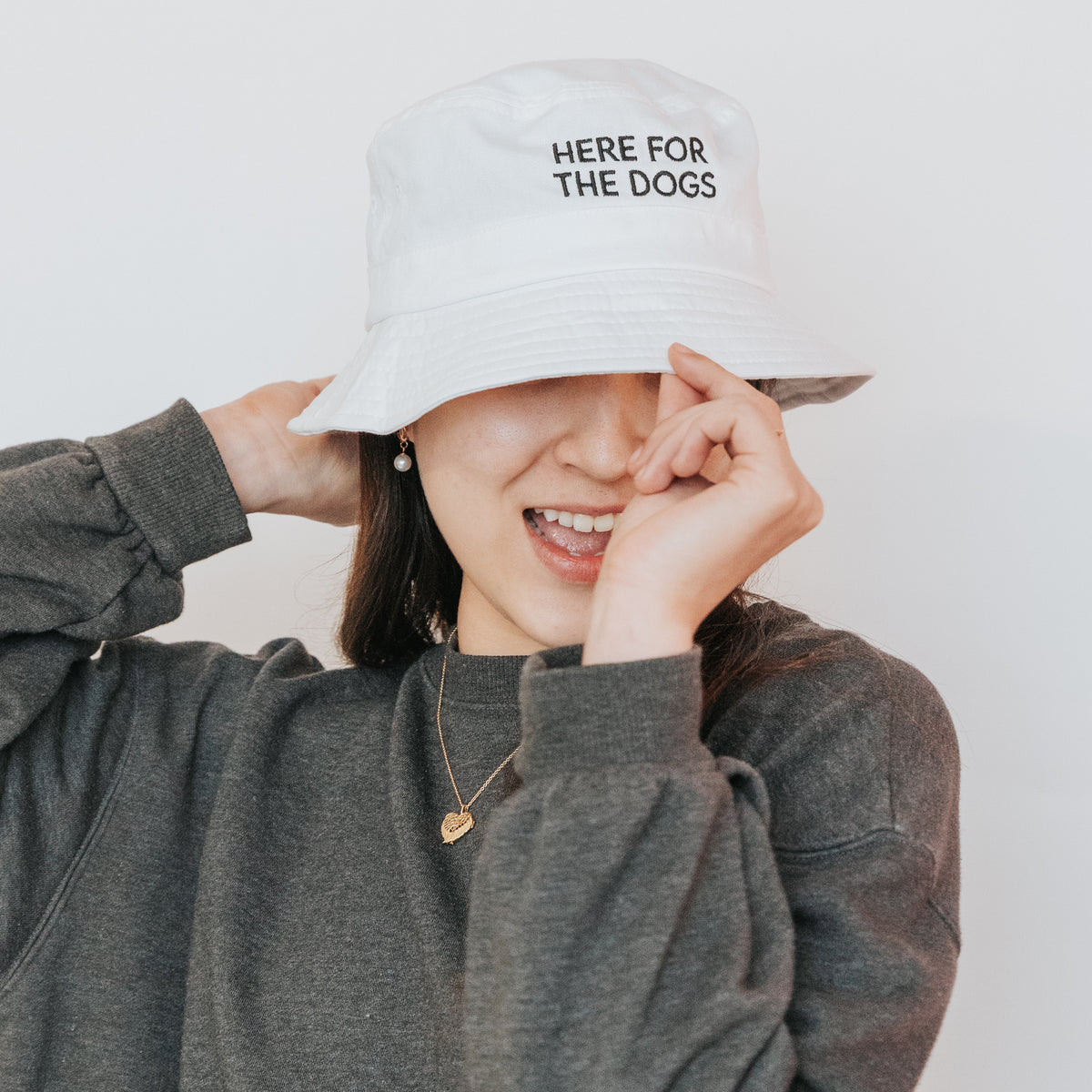 A smiling person in a gray sweater and necklace adjusts the Sassy Woof Bucket Hat - Here For the Dogs, which partially covers their eyes. The white hat features "HERE FOR THE DOGS" text. The background is plain white.