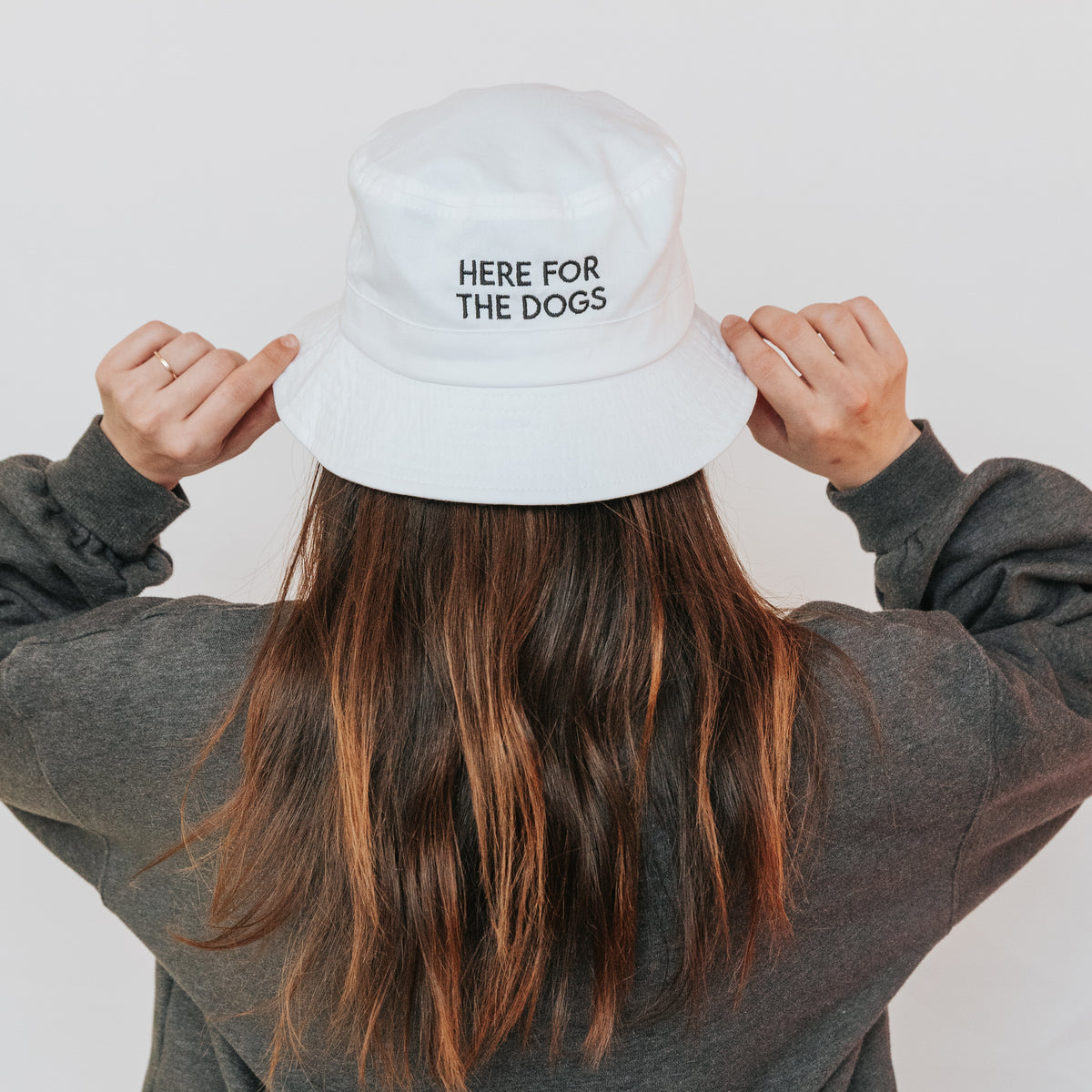 A person with long brown hair, facing away from the camera, wears a white Sassy Woof "Here For the Dogs" embroidered bucket hat and a dark sweatshirt, holding the brim of the hat with both hands.