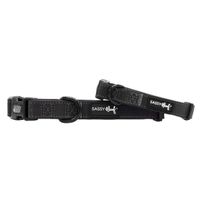 Two black Sassy Woof "Dog Collar - Reflection" adjustable collars with metal rings and buckles, white stitching, logo tags, and reflective features for nighttime safety are displayed on a white background.