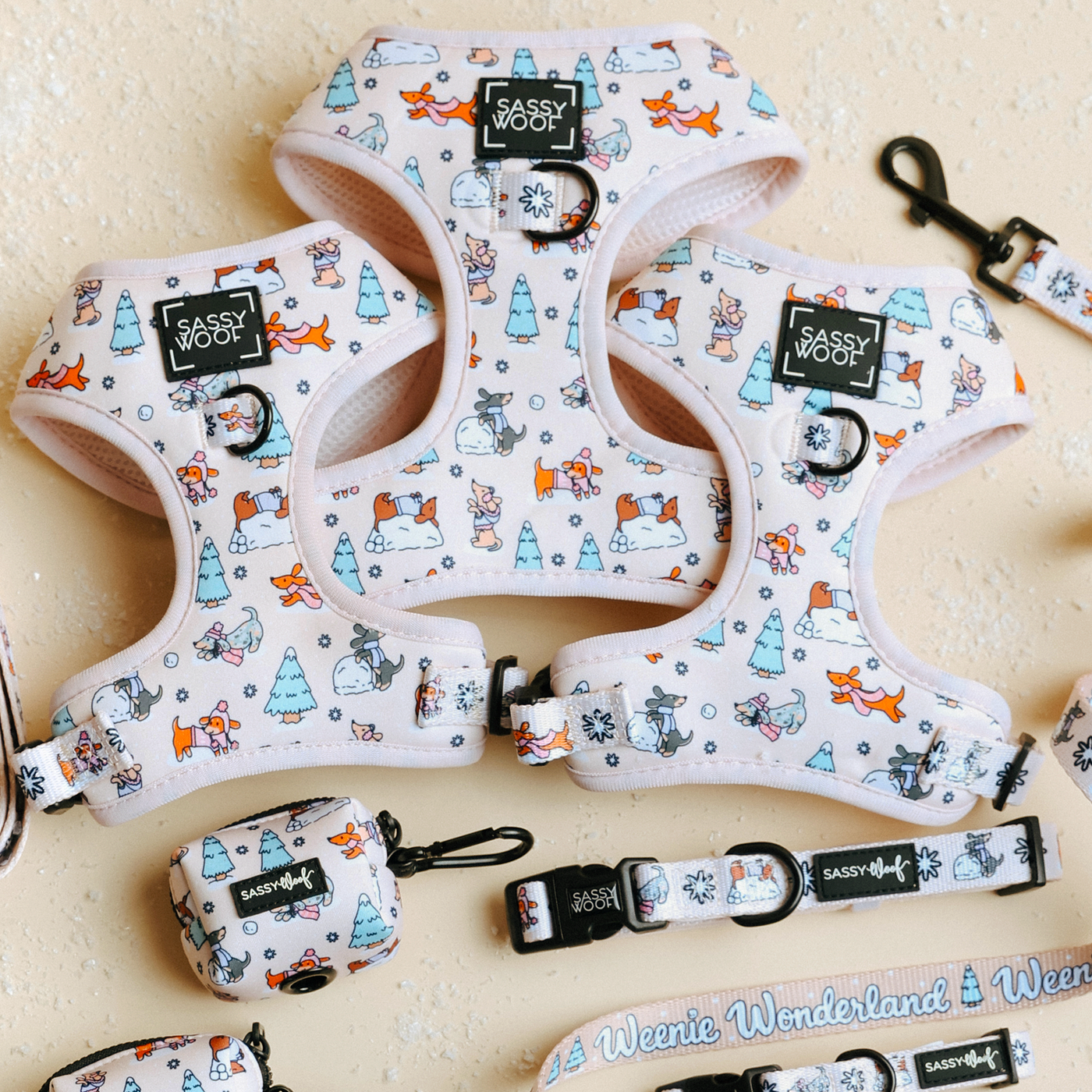 Three Sassy Woof Dog Four Piece Bundles from the Weenie Wonderland Collection are displayed on a light textured background. Each bundle includes a pastel pink adjustable harness with whimsical cartoon dog and winter tree patterns, a matching collar, leash, and small bag holder—perfectly crafted for both comfort and style as an ideal dachshund harness.