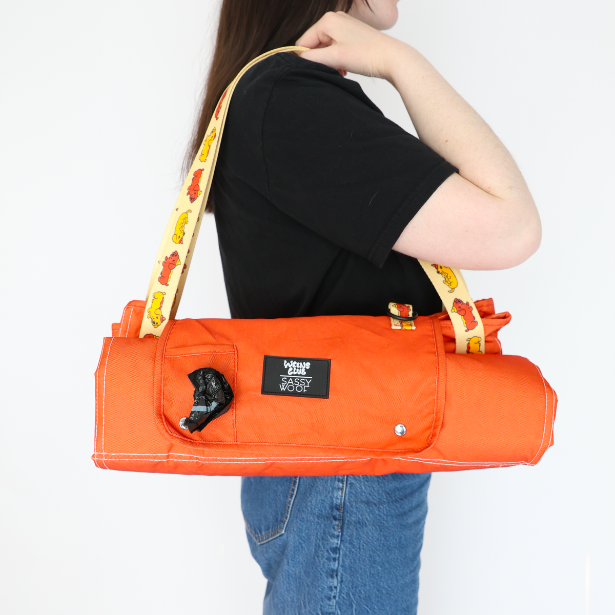 A person with long brown hair carries the Sassy Woof Dog Picnic Blanket - Hot Dawg, featuring an orange design inspired by dachshunds, a yellow strap with dog illustrations, a black label, and a built-in dispenser for waste bags.