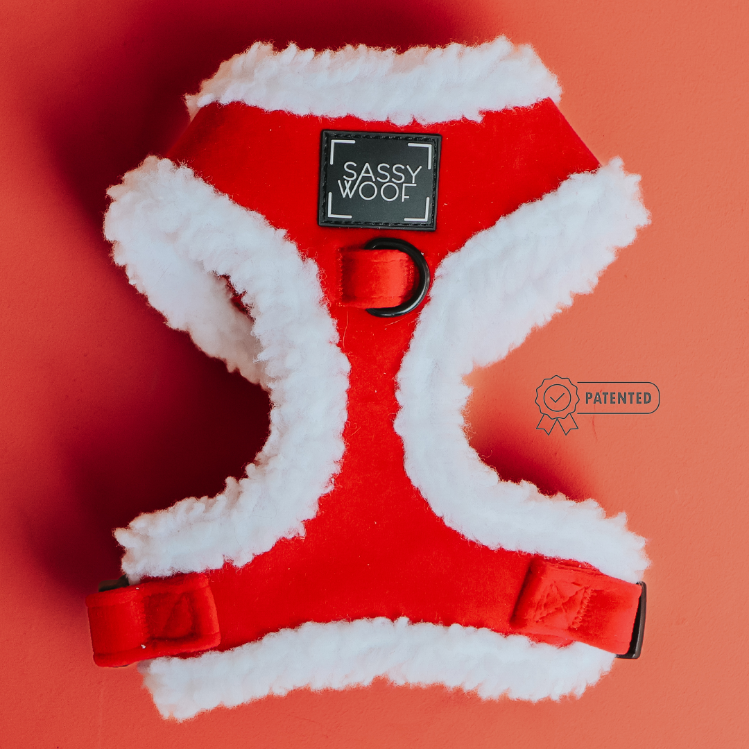 The Dog Adjustable Harness - Naughty by Sassy Woof, in red and white sherpa with a black label, is shown on a coral background—ideal for cozy holiday looks. A small Patented badge icon is visible beside the harness.