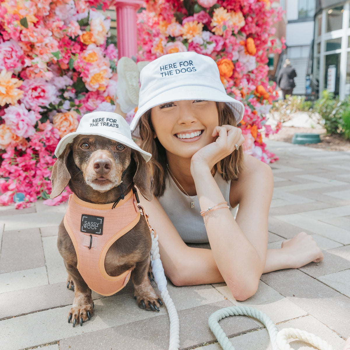 A smiling woman and her brown dog wear matching Sassy Woof bucket hats from the "Here For The Treats" Bundle, posing before vibrant pink and orange flowers. The dog's vest reads "Sassy & Gassy," adding extra charm to their playful style.
