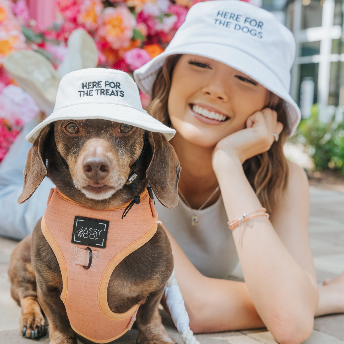 A smiling woman and her brown dachshund wear the Sassy Woof Bucket Hat Bundle - Here For The Treats, each in matching hats. Flowers bloom brightly in the background.