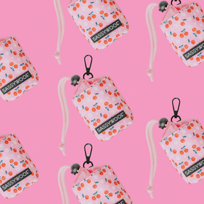 A repeating pattern of Sassy Woof's Mon Chérry Treat Pouch and matching pink dog poop bag holders with cherry prints and black clips, all showcased on a bright pink background.