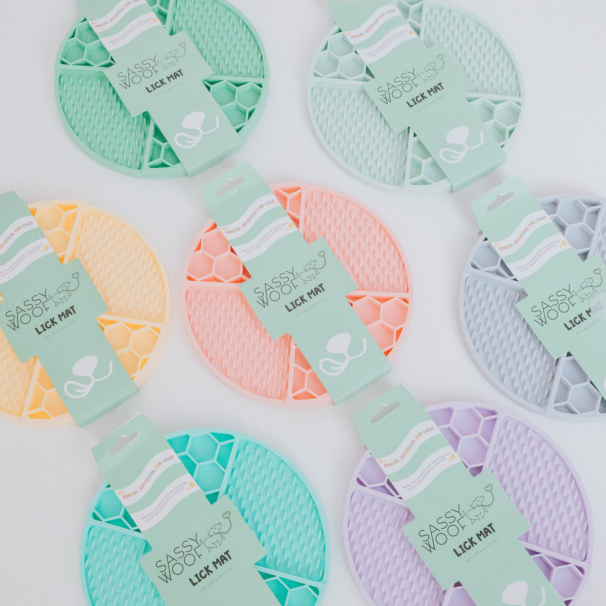 Six pastel Sassy Woof Dog Lick Mats—yellow, green, pink, blue, purple, and orange—are arranged in two rows. Each mat displays the Sassy Woof name plus coffee cup and bone illustrations.