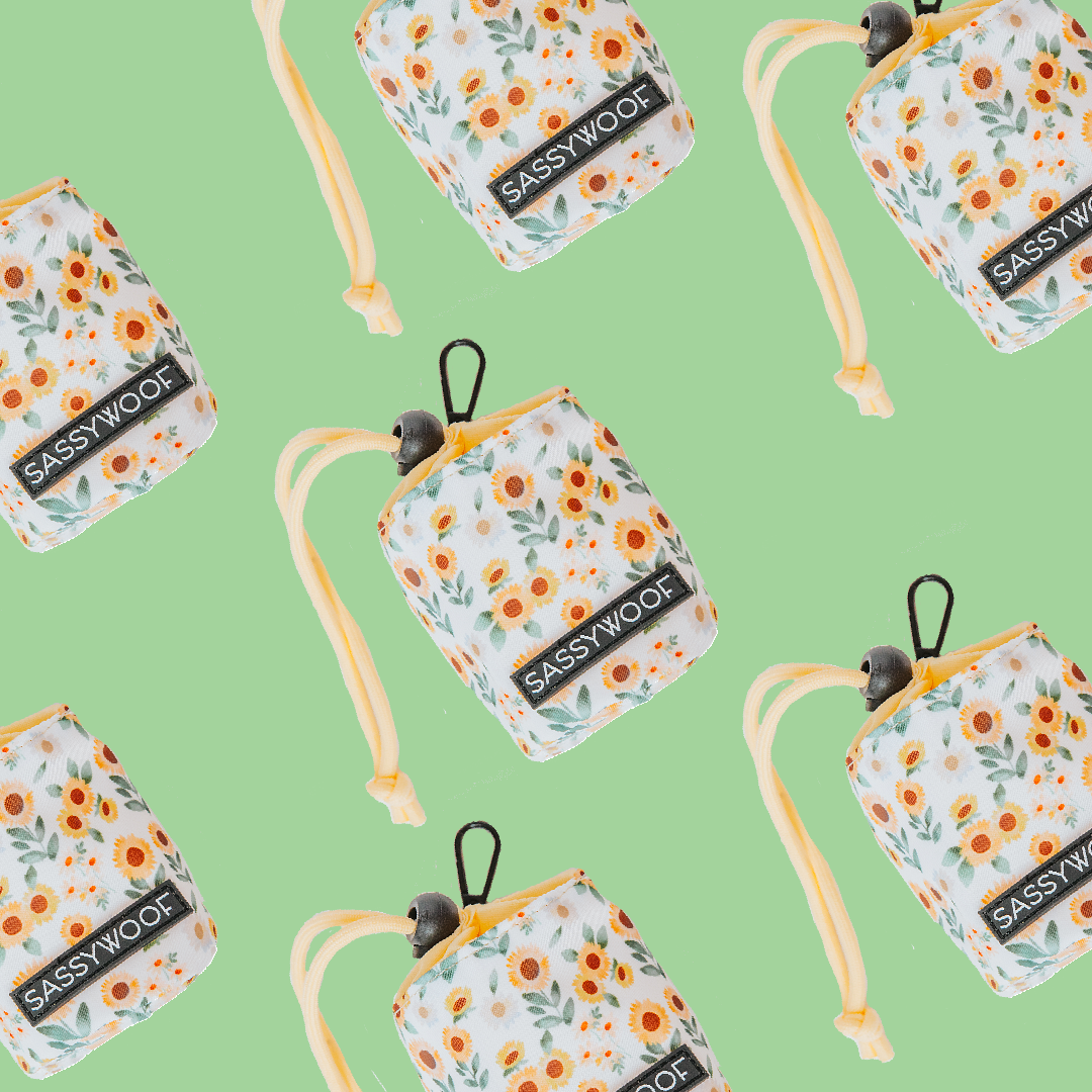 The Sassy Woof “Treat Pouch - Sunflower Dreams” features a white floral design with orange flowers, green leaves, and yellow straps on a light green background—stylish and perfect for pet owners.
