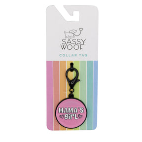The Dog Collar Tag - Mama's Girl by Sassy Woof is a round pink pet tag with "Mamas Girl" in white and black, attached to black hardware, and comes on pastel rainbow-striped Sassy Woof packaging.