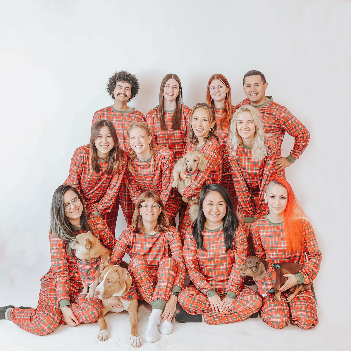 Thirteen people in Sassy Woof's "Deck the Paws" pajamas pose together, smiling with four dogs in front of them, all against a plain white background.