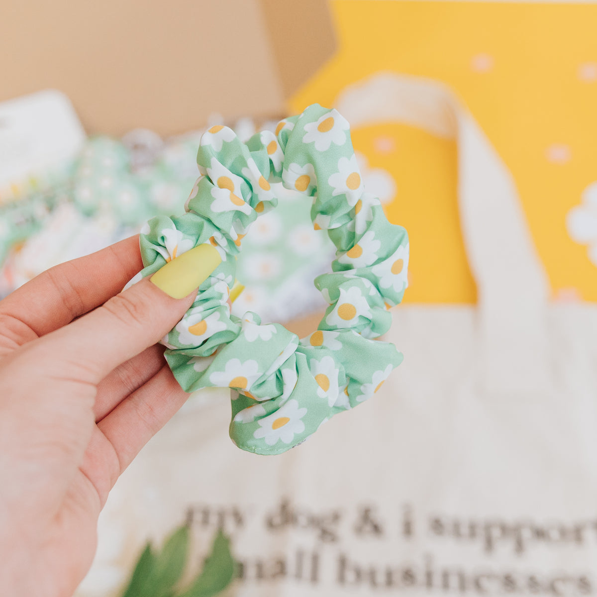 A hand with yellow-painted nails holds the Sassy Woof Daisy Scrunchie, adorned with white and yellow daisy flowers. In the background, a white tote bag and a yellow daisy-patterned surface are visible—great for pairing with matching dog accessories.