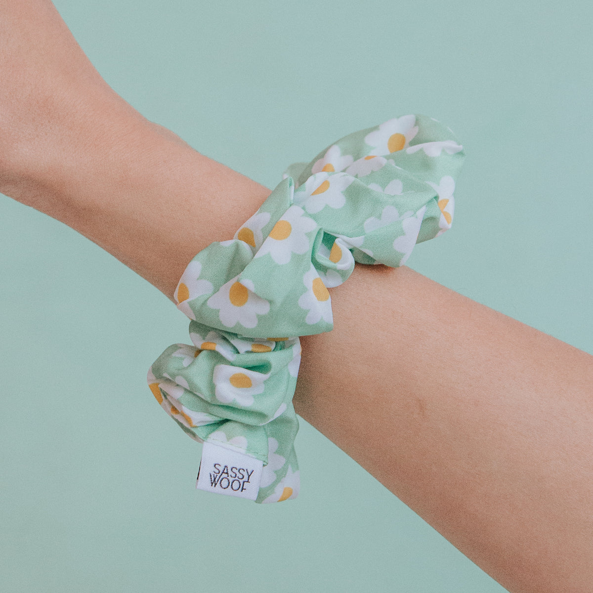 A person’s arm with a Sassy Woof Daisy Scrunchie—featuring white and yellow daisies and a “Sassy Woof” label—wrapped around their wrist against a matching light green background, ideal for coordinating with dog accessories.