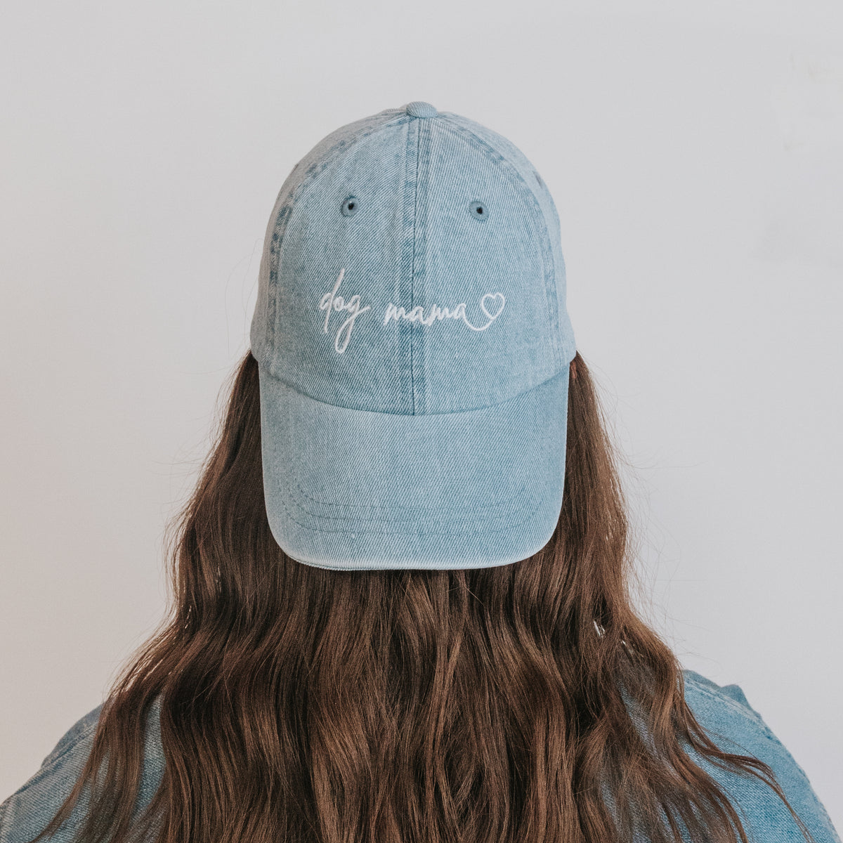 A person with long brown hair, seen from behind, wears the Sassy Woof Dog Mama Hat - Denim—a light blue embroidered cap with a heart motif on the front—against a plain white background.