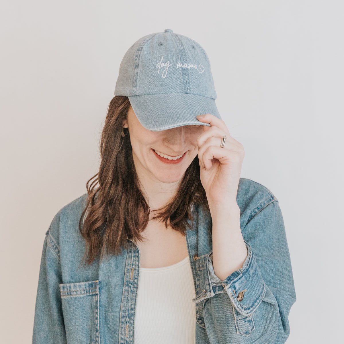 A woman with wavy brown hair smiles, tilting her head and holding the brim of her Sassy Woof Dog Mama Hat - Denim while wearing a light denim jacket.