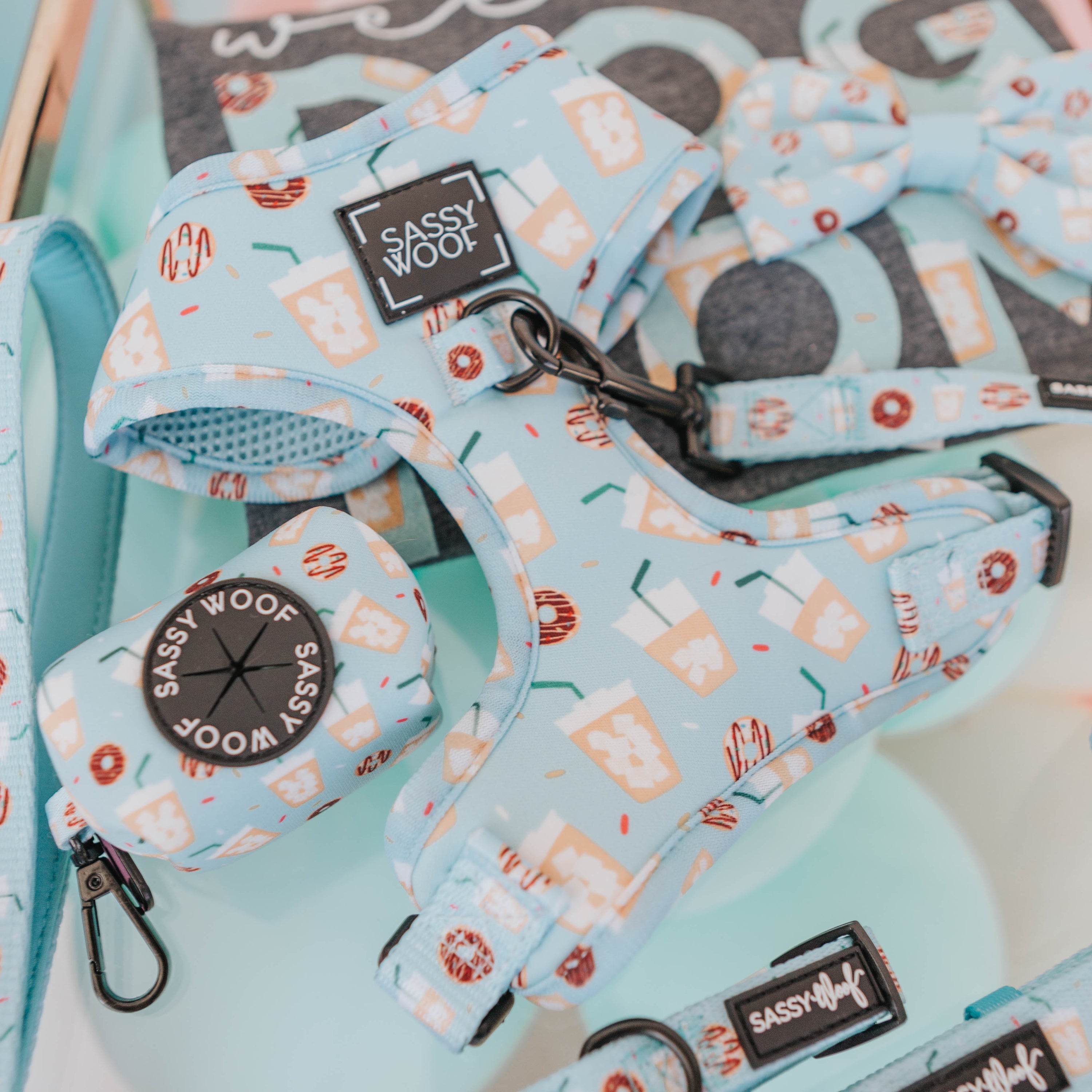 The Sassy Woof Dog Four Piece Bundle - Sweets & Pawfee, featuring light blue harness and accessories with food and drink designs, is displayed on a pastel background with a matching leash and part of a sign or card visible above.