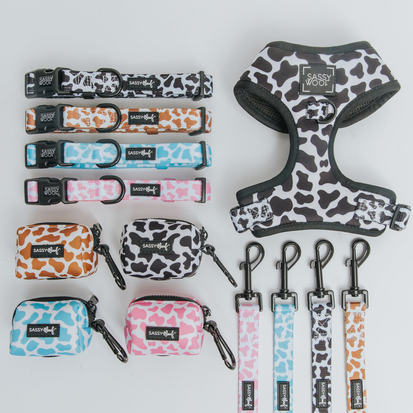 Dog accessories set with cow print designs in black, brown, pink, and blue. Includes collars, leashes, harness, and waste bag holders, all labeled “Sassy Woof,” arranged neatly on a white background.