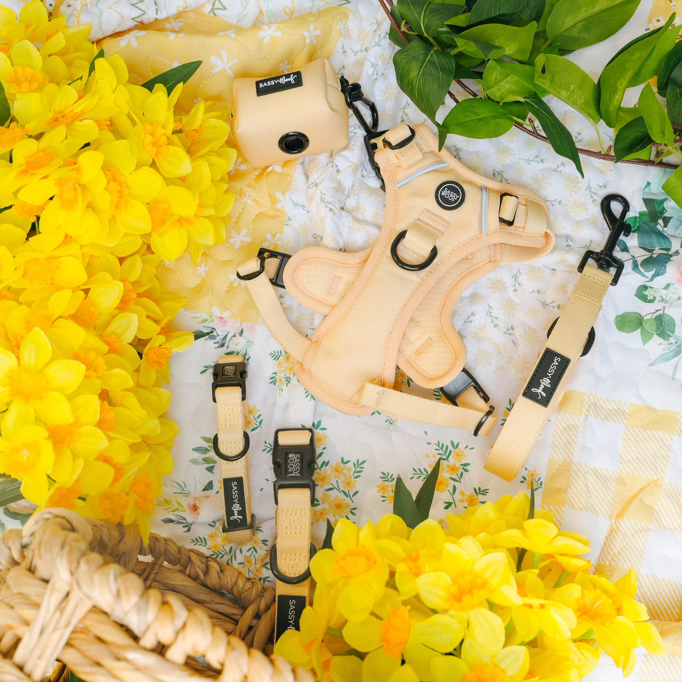 A pastel yellow dog harness, leash, and collar set displayed on a floral picnic blanket with yellow flowers, green leaves, and a wicker basket.