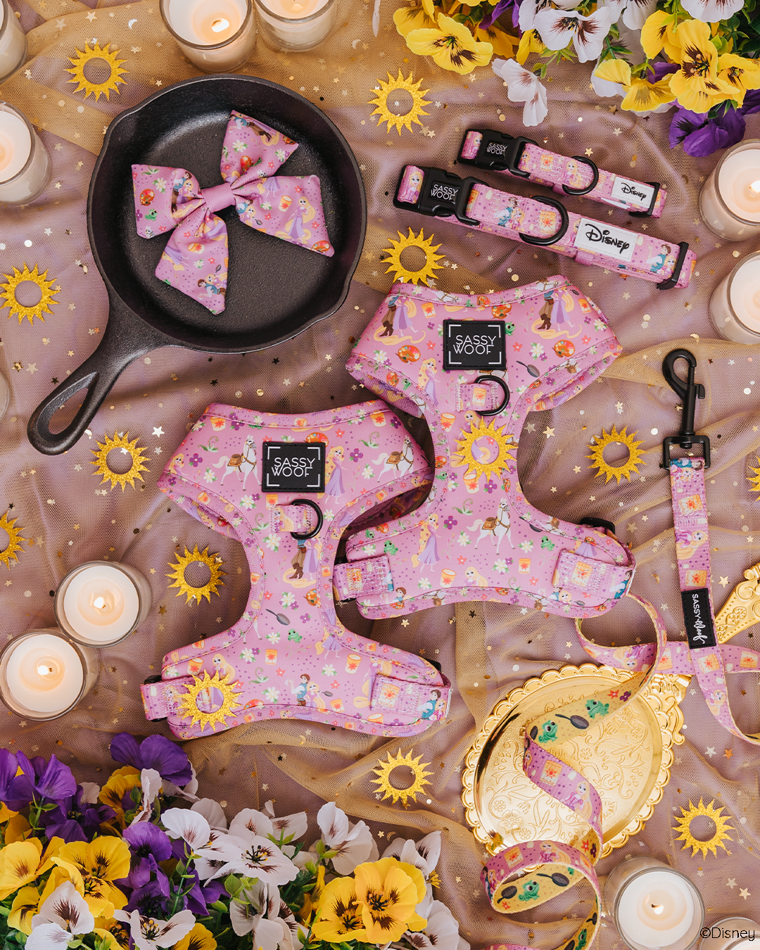 Flat lay of Rapunzel-themed dog accessories on a gold mesh fabric, surrounded by flowers, candles, a frying pan, a golden mirror, and a spool of ribbon. The accessories are pink with Disney motifs.