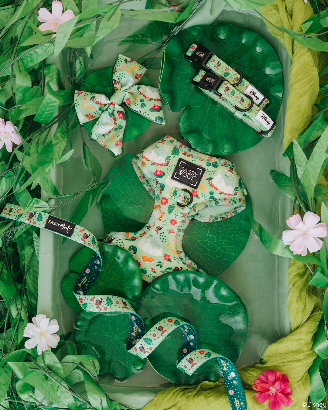A set of green and floral-patterned pet accessories, including a harness, collars, bow tie, and leash, arranged on faux lily pads and surrounded by green leaves and pink flowers.