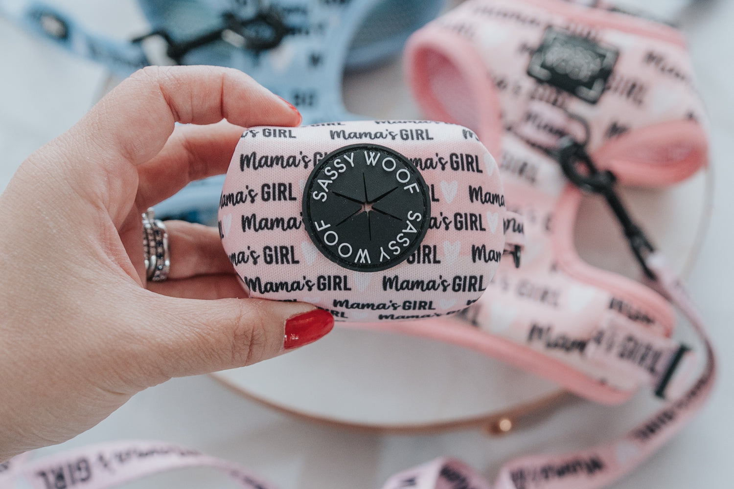 A hand with red-painted nails holds a pink dog accessory labeled Mamas Girl and Sassy Woof, with matching pink and blue dog harnesses and leashes in the background.