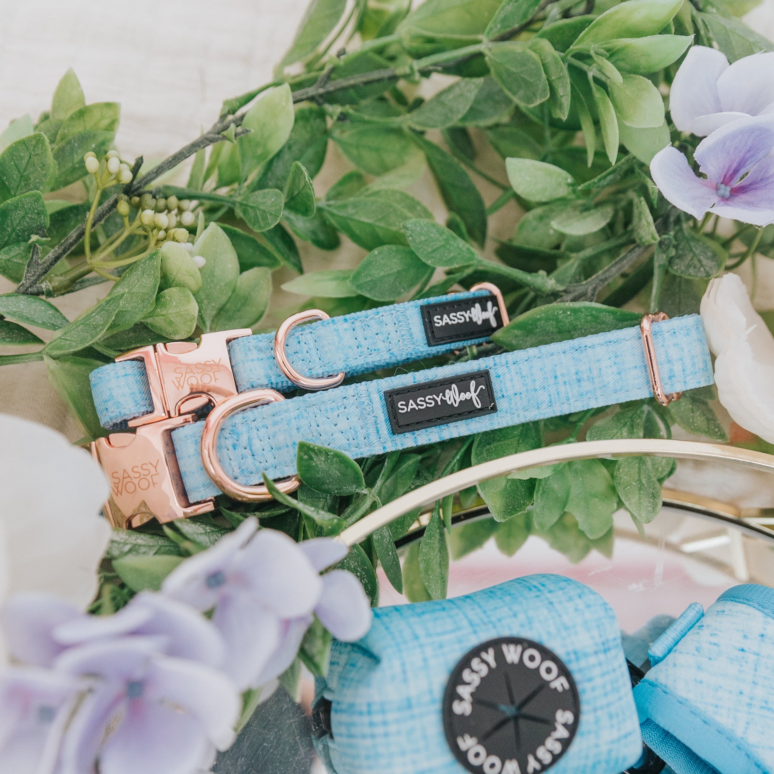 A Dog Collar Three Piece Bundle - Blumond by Sassy Woof, featuring light blue collars with rose gold hardware and a coordinating leash, is artfully arranged amid green leaves and purple flowers for a fresh and stylish pet accessory display.