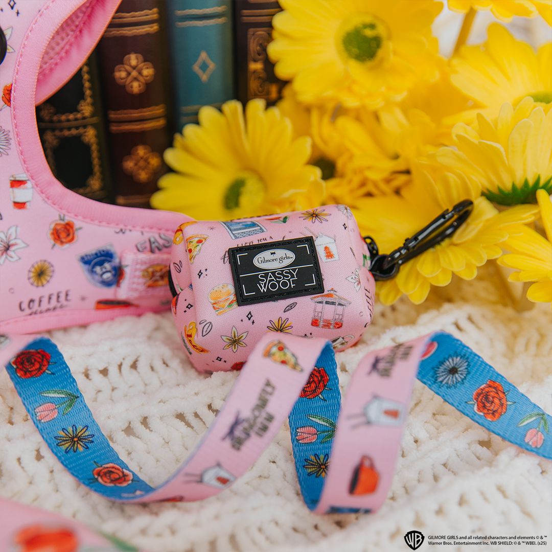 Two blue Sassy Woof dog collars with rose gold hardware are displayed among green leaves and purple flowers. The Sassy Woof label is visible on each collar.