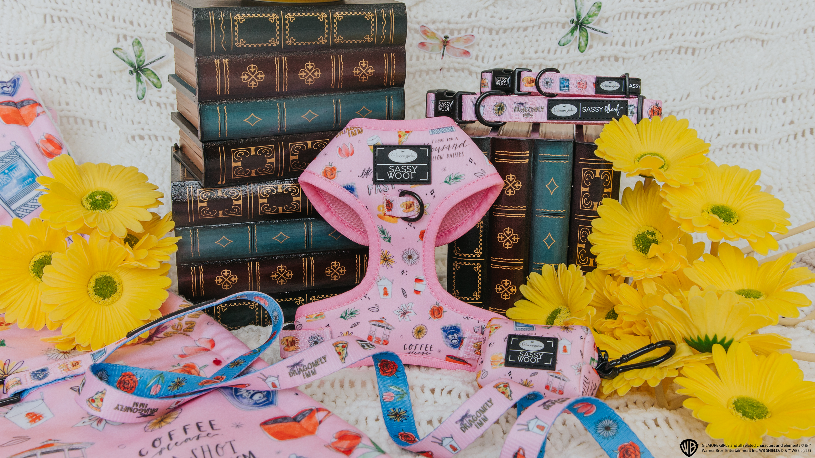 A pink dog harness, leash, and collars with coffee-themed prints are displayed among vintage books and bright yellow daisies on a white crochet backdrop.