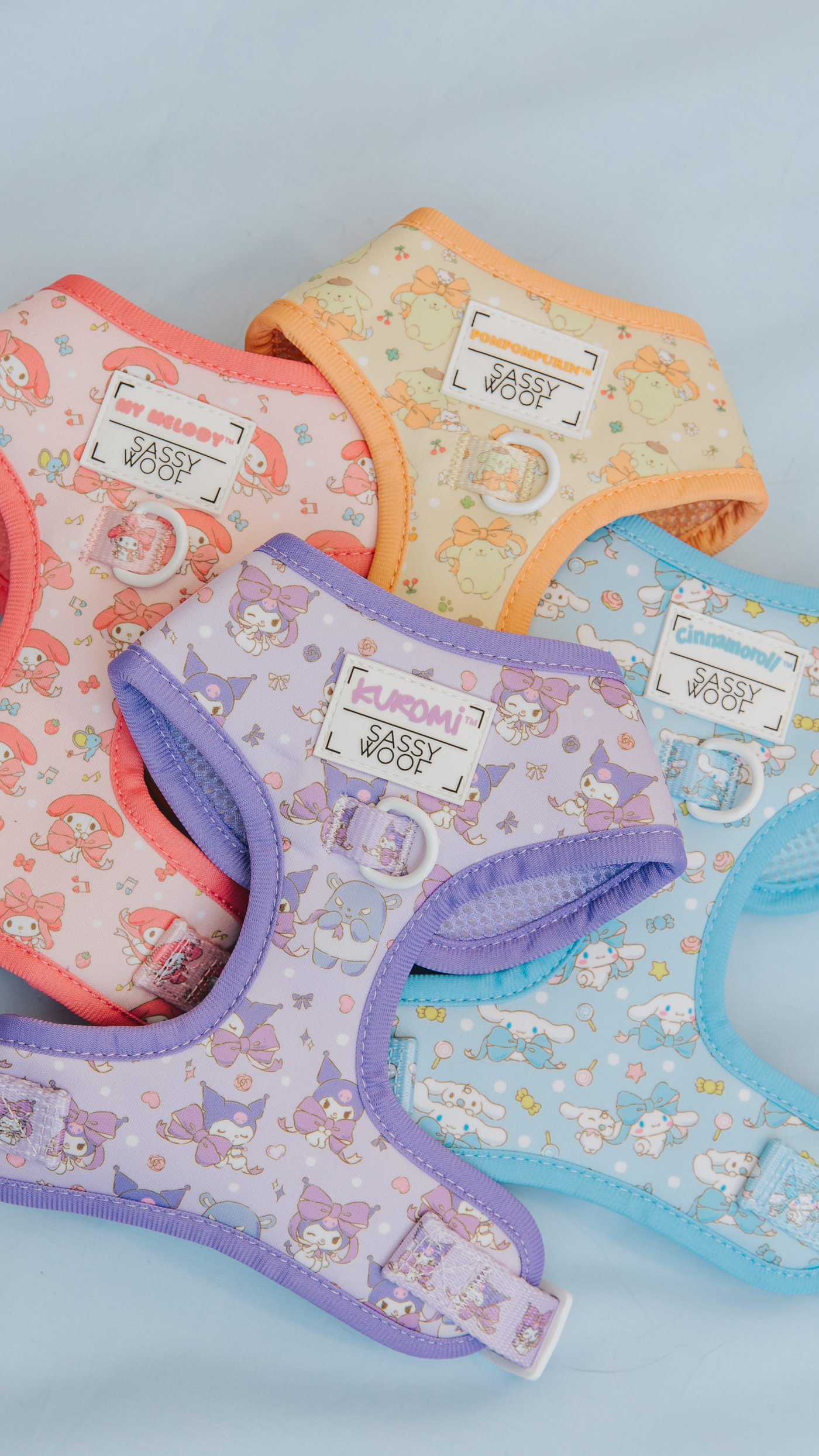 Four colorful pet harnesses with cute cartoon patterns are laid out on a light blue surface. The harnesses are pink, orange, purple, and blue, each featuring playful character designs.