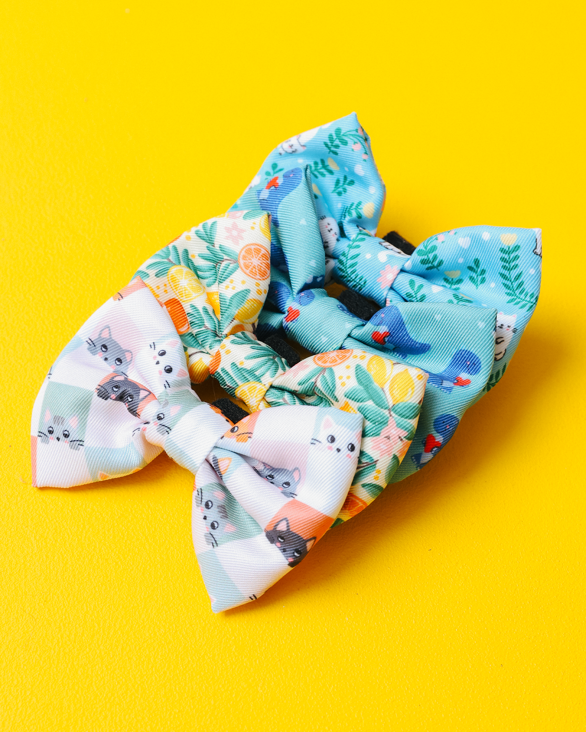 A hand with white-painted nails holds a colorful, citrus-patterned bow in front of a green leafy wreath decorated with lemons and white flowers on a bright yellow background.