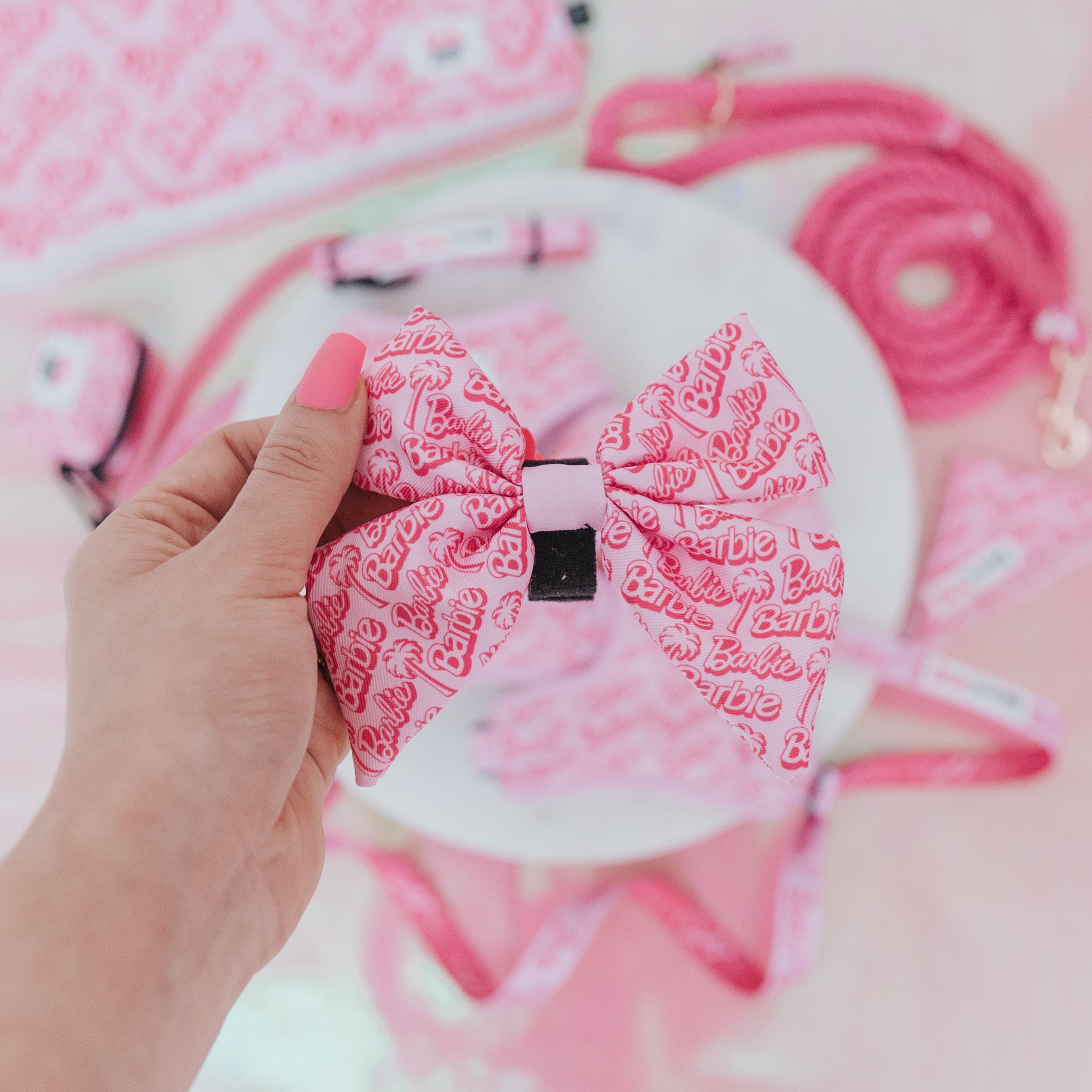 A hand with pink-painted nails holds the Dog Sailor Bow - BARBIE™ Malibu by Sassy Woof. The background showcases matching pink dog accessories from the Barbie x Sassy Woof collection, including an adjustable harness, leash, and tags.