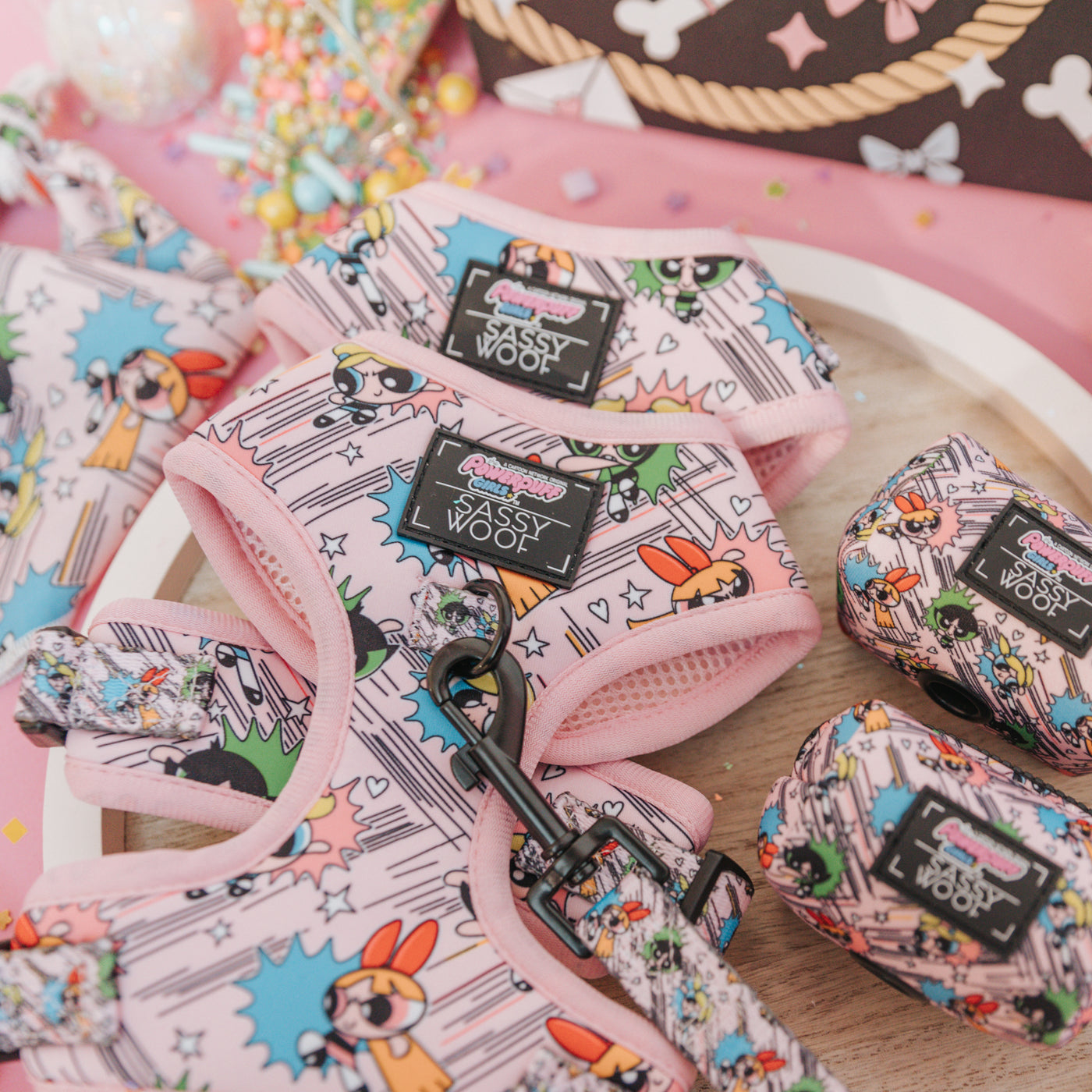 A close-up of pink dog harnesses, leashes, and accessories featuring colorful Powerpuff Girls designs, laid out on a wooden tray with pastel decorations around them.