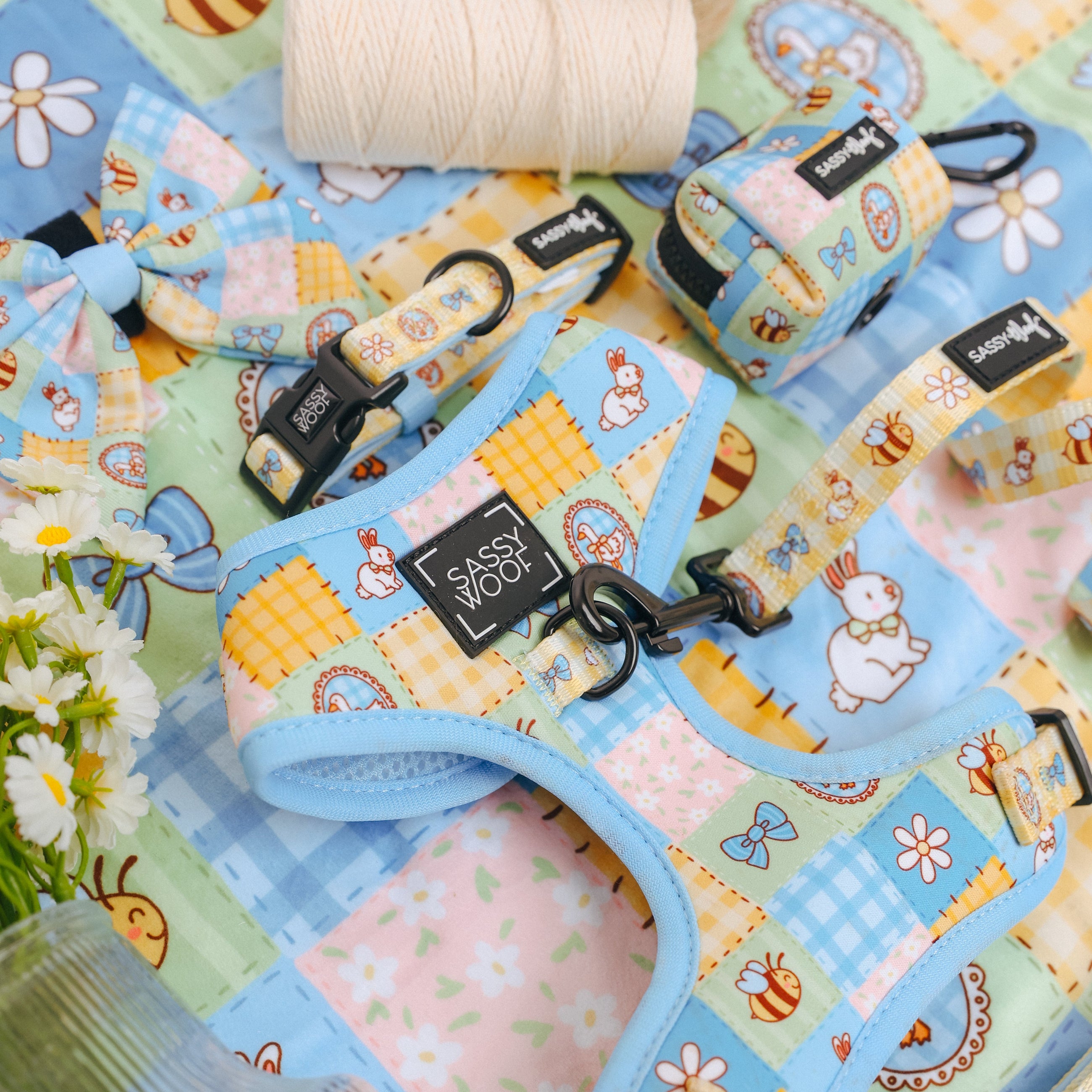 Colorful pet harness, collar, leash, and bowtie with cartoon animal prints lie on a pastel patchwork quilt. Nearby are white daisies, a spool of string, and a glass jar.