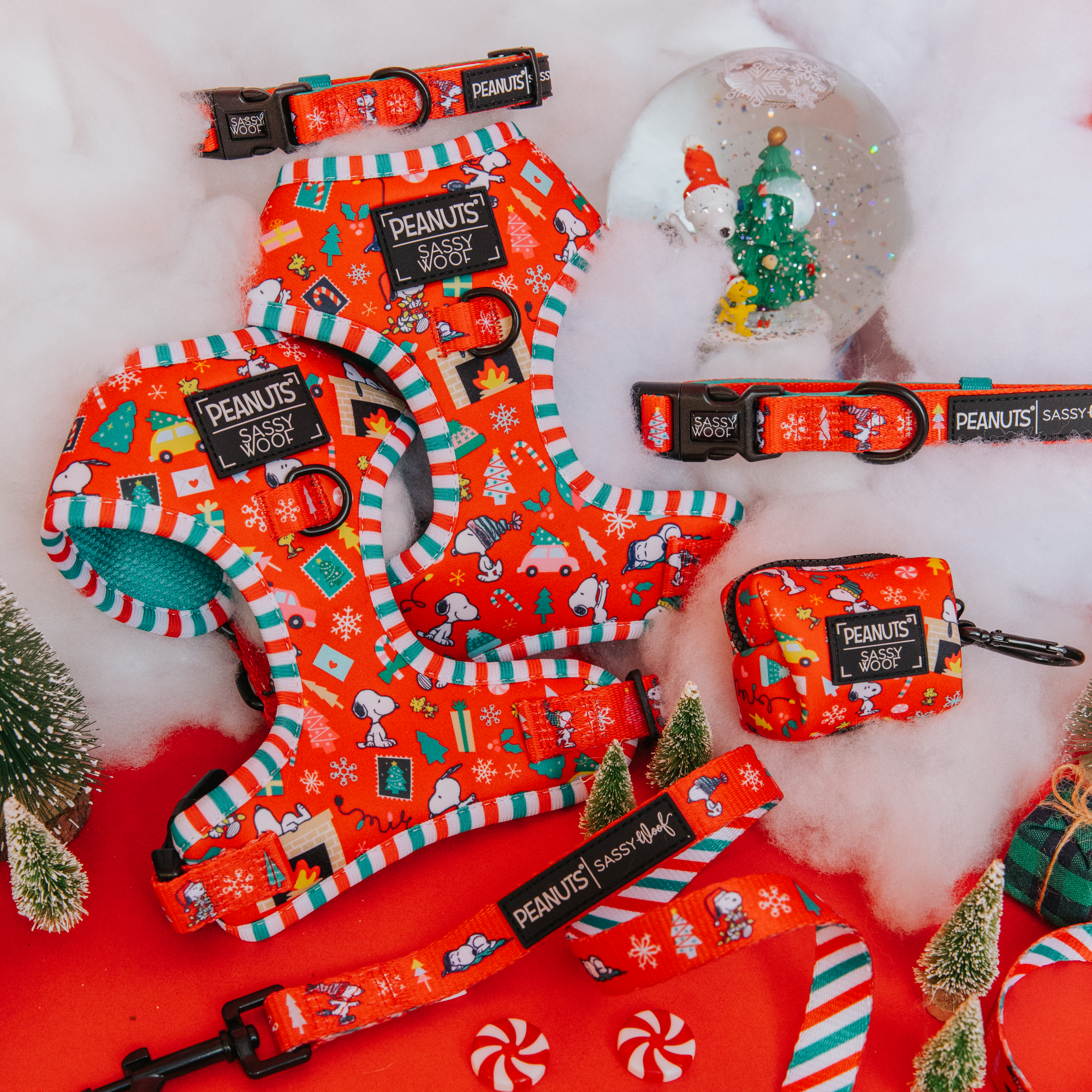 A set of red dog accessories with a festive Peanuts Christmas design, including harnesses, collars, a leash, and a poop bag holder, displayed on a red background with holiday decorations and fake snow.