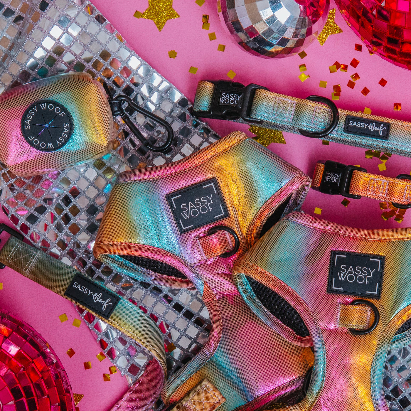 Iridescent dog harnesses, collars, and a waste bag holder labeled Sassy Woof are arranged on a pink background with disco balls, confetti, and a silver sequin cloth nearby.