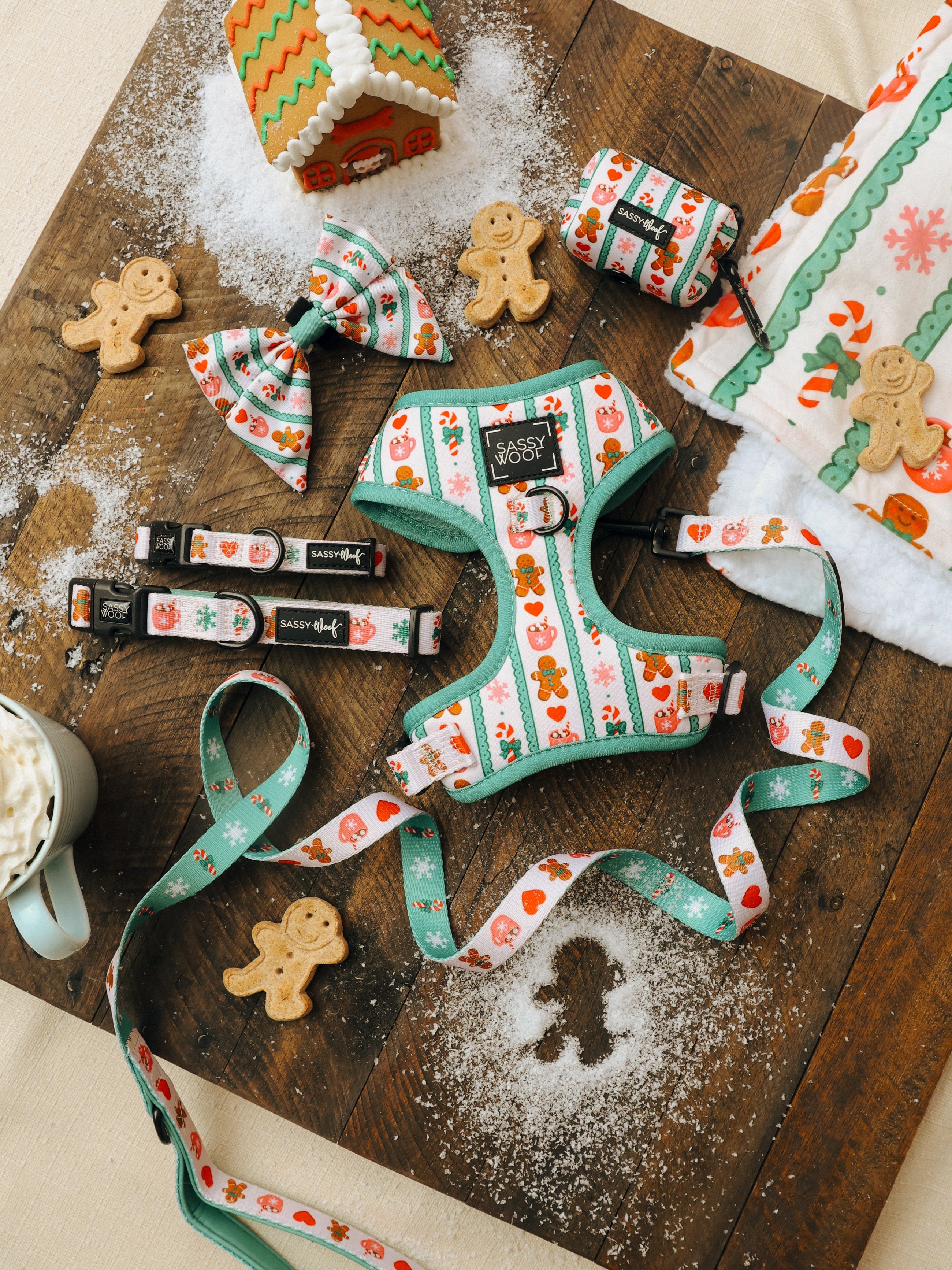 Dog harness, collar, leash, bow tie, and waste bag holder with gingerbread patterns on a dusted wooden board, surrounded by gingerbread cookies, a mug of cocoa, and festive linens.