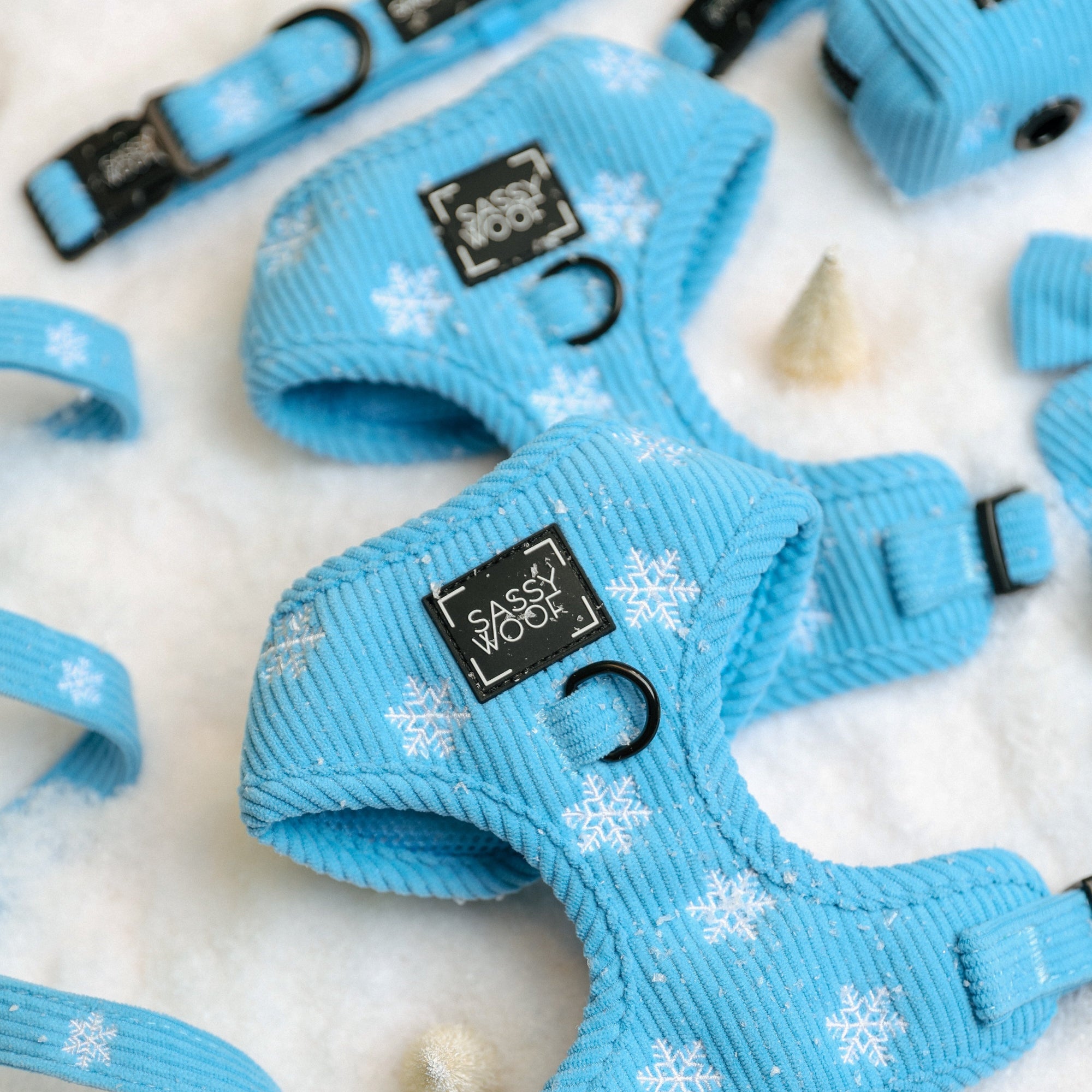 Sassy Woof Dog Four Piece Bundle - Morning Frost featuring a light blue adjustable harness, collar, leash, and waste bag holder with a white snowflake pattern, arranged on a white snowy surface.